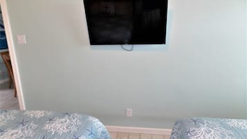 Habitación con televisor de pantalla plana montado en la pared, dos camas con ropa de cama azul y piso de baldosas.