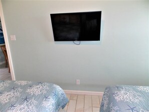Habitación con televisor de pantalla plana montado en la pared, dos camas con ropa de cama azul y piso de baldosas.