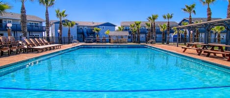 A clear blue pool surrounded by lounge chairs and palm trees, with buildings in the background.