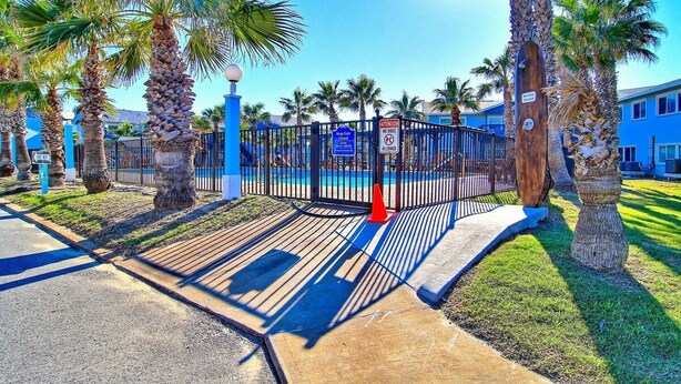 A gated entrance with palm trees, a pool, and a blue building in the background. - Captains Quarters Large Pool Boardwalk to Beach (Port Aransas)