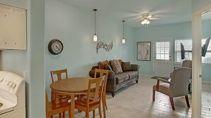 A dining area with a round table and chairs, a sofa with cushions, a ceiling fan, and a wall-mounted clock. - Captains Quarters Large Pool Boardwalk to Beach (Port Aransas)