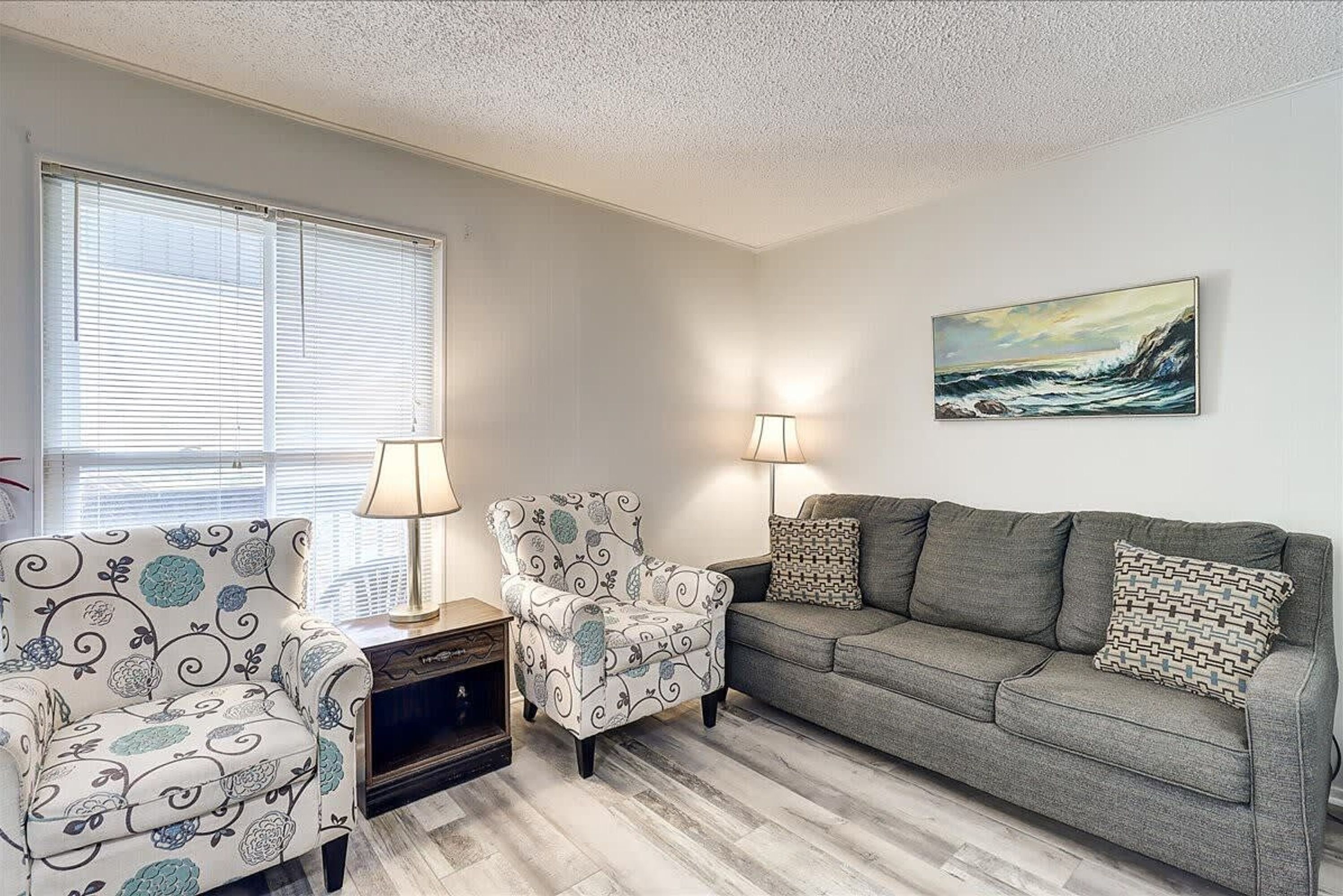 A living room with a grey sofa, two patterned armchairs, a side table, and a painting on the wall.