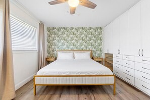 Une chambre à coucher comprenant un lit, un ventilateur de plafond, une table de chevet, une armoire et une fenêtre avec des stores.