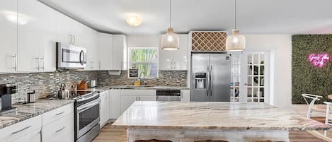 A modern kitchen with white cabinetry, a large island with stools, stainless steel appliances, and a neon sign.
