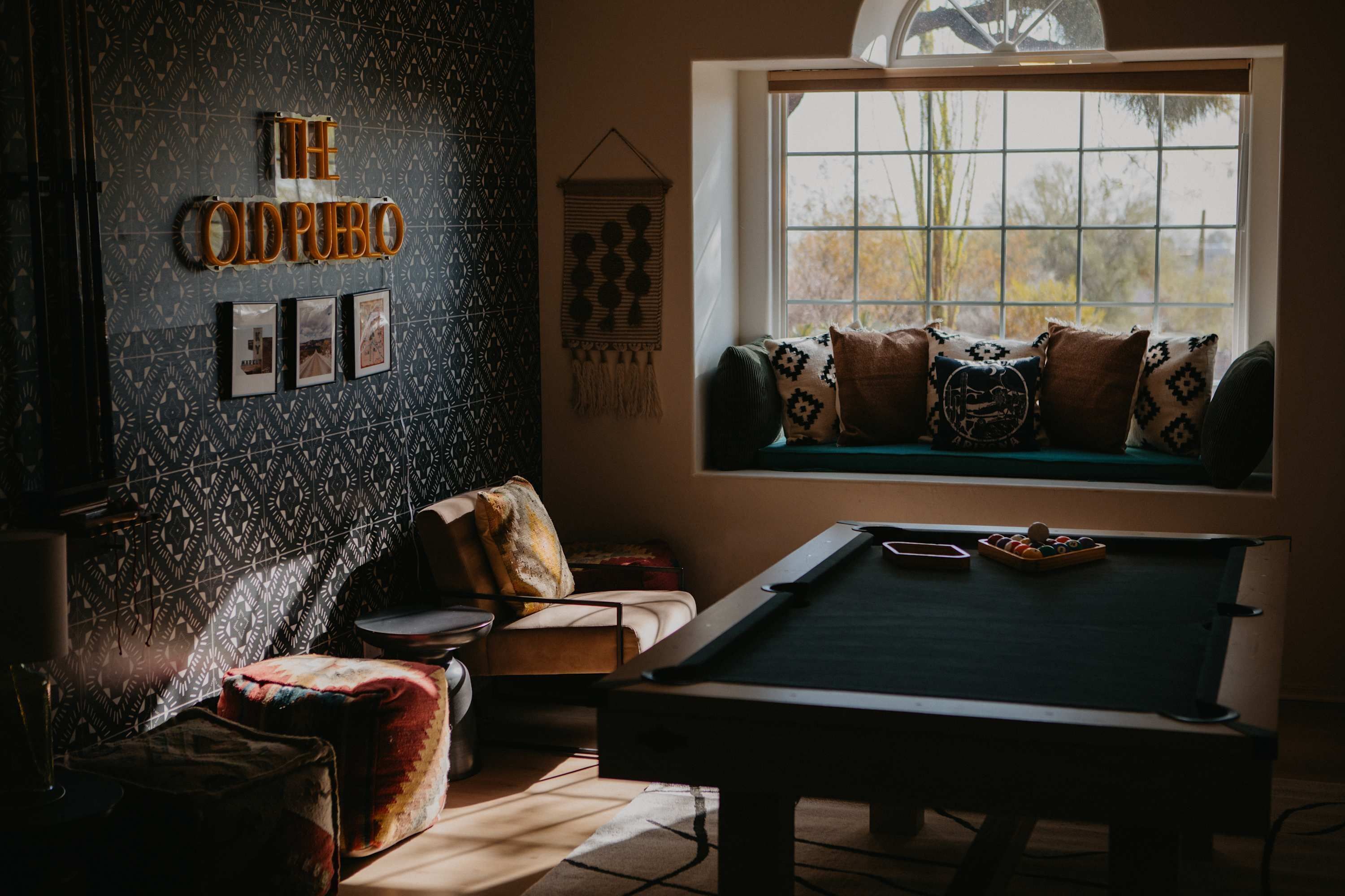 Una habitación con mesa de billar, sofá con cojines estampados, pared con diseño geométrico y ventana con vista a árboles.