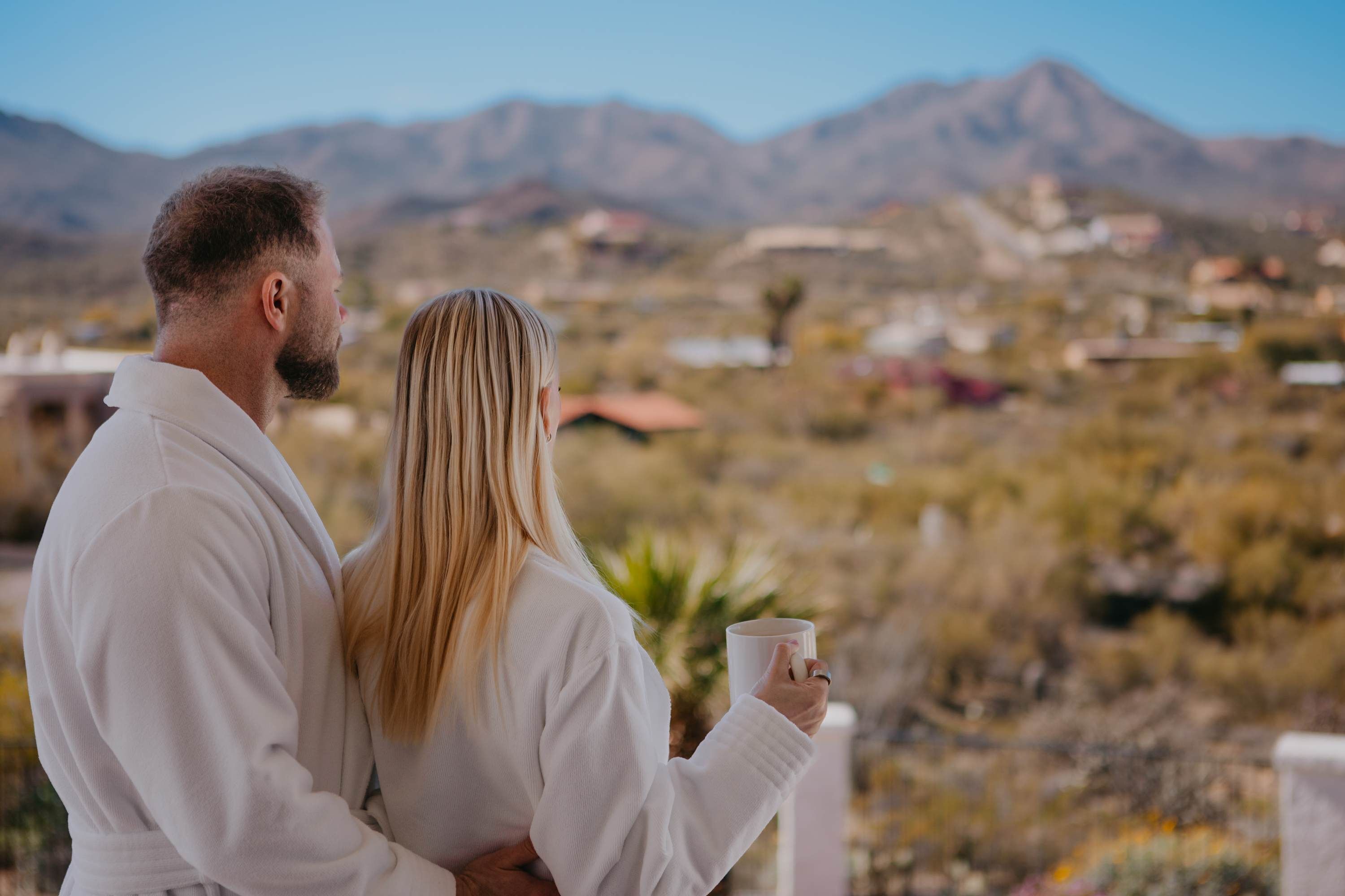 Una pareja está de pie en un balcón con vistas a un paisaje montañoso y edificios a lo lejos.