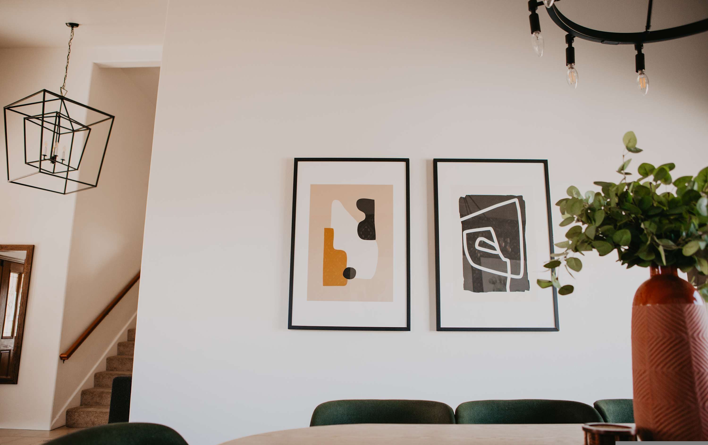 Un espace repas avec un escalier, un luminaire suspendu et deux œuvres d’art abstraites encadrées accrochées au mur.