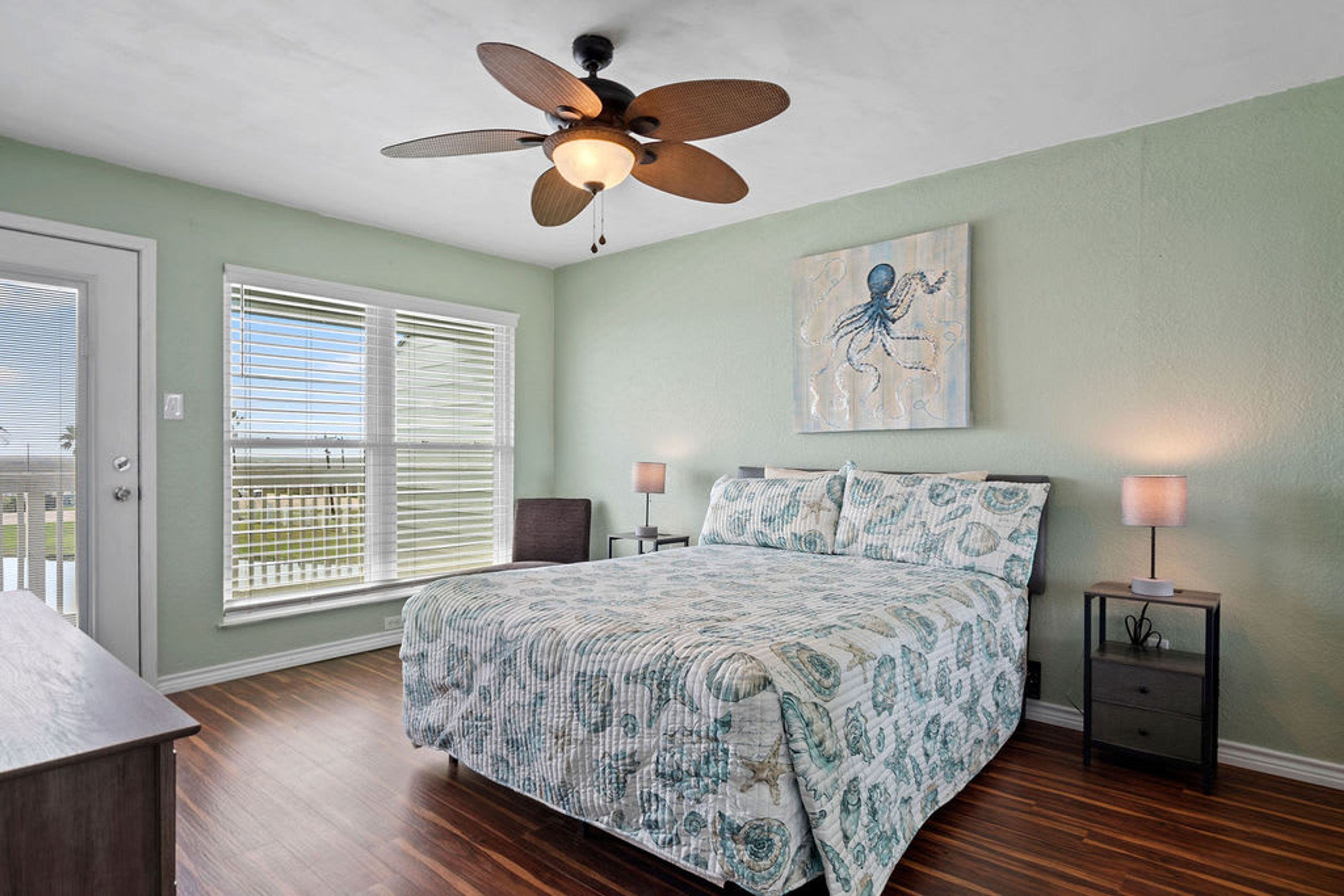 A bedroom with a bed, a ceiling fan, a window with blinds, a nightstand, and a wall-mounted painting.