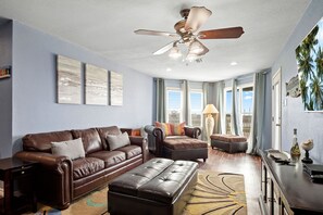 Ein Wohnzimmer mit braunem Ledersofa, passendem Hocker, Deckenventilator und Blick ins Freie durch große Fenster.