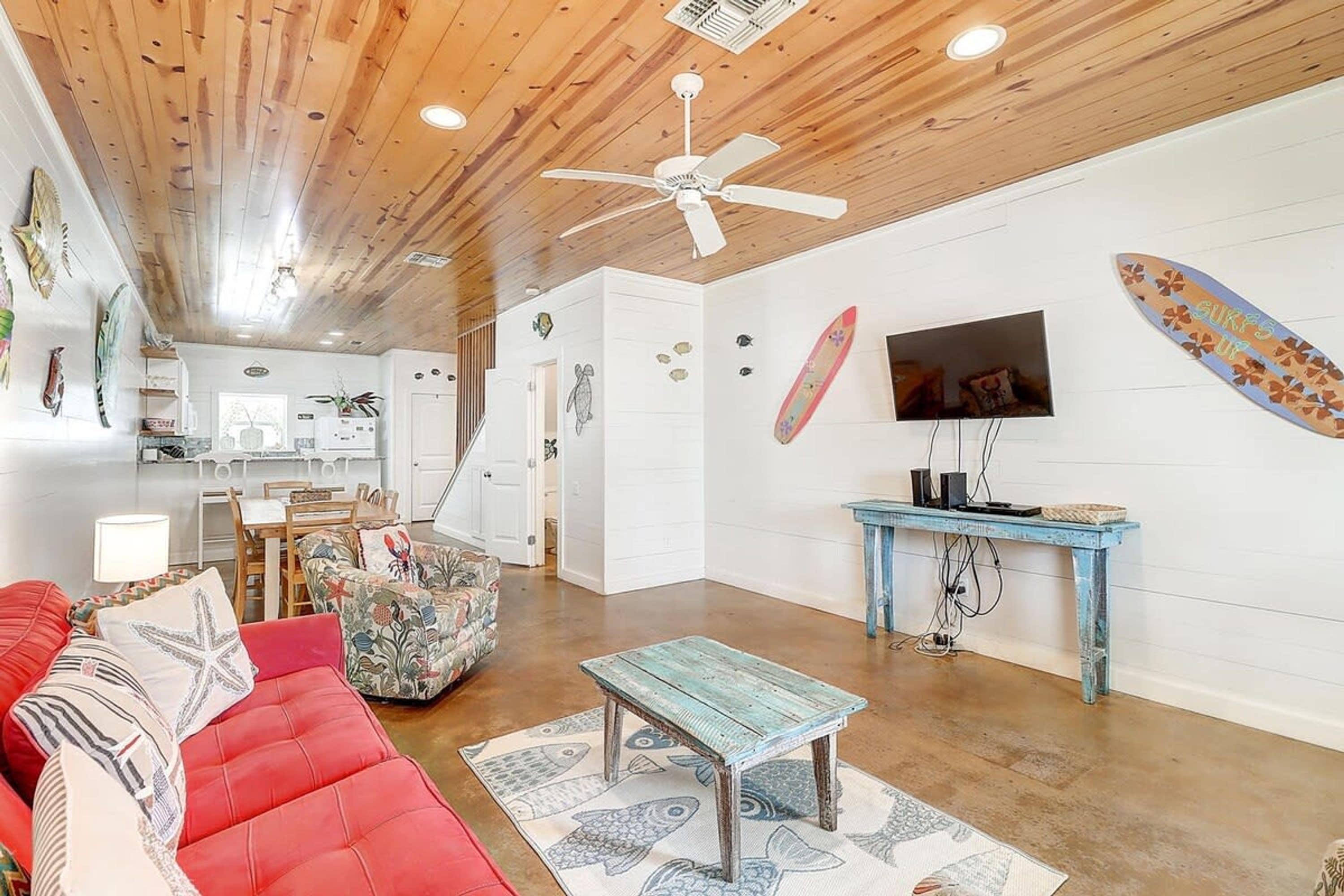 A living room with a red sofa, a blue coffee table, a ceiling fan, and a surfboard decoration on the wall.