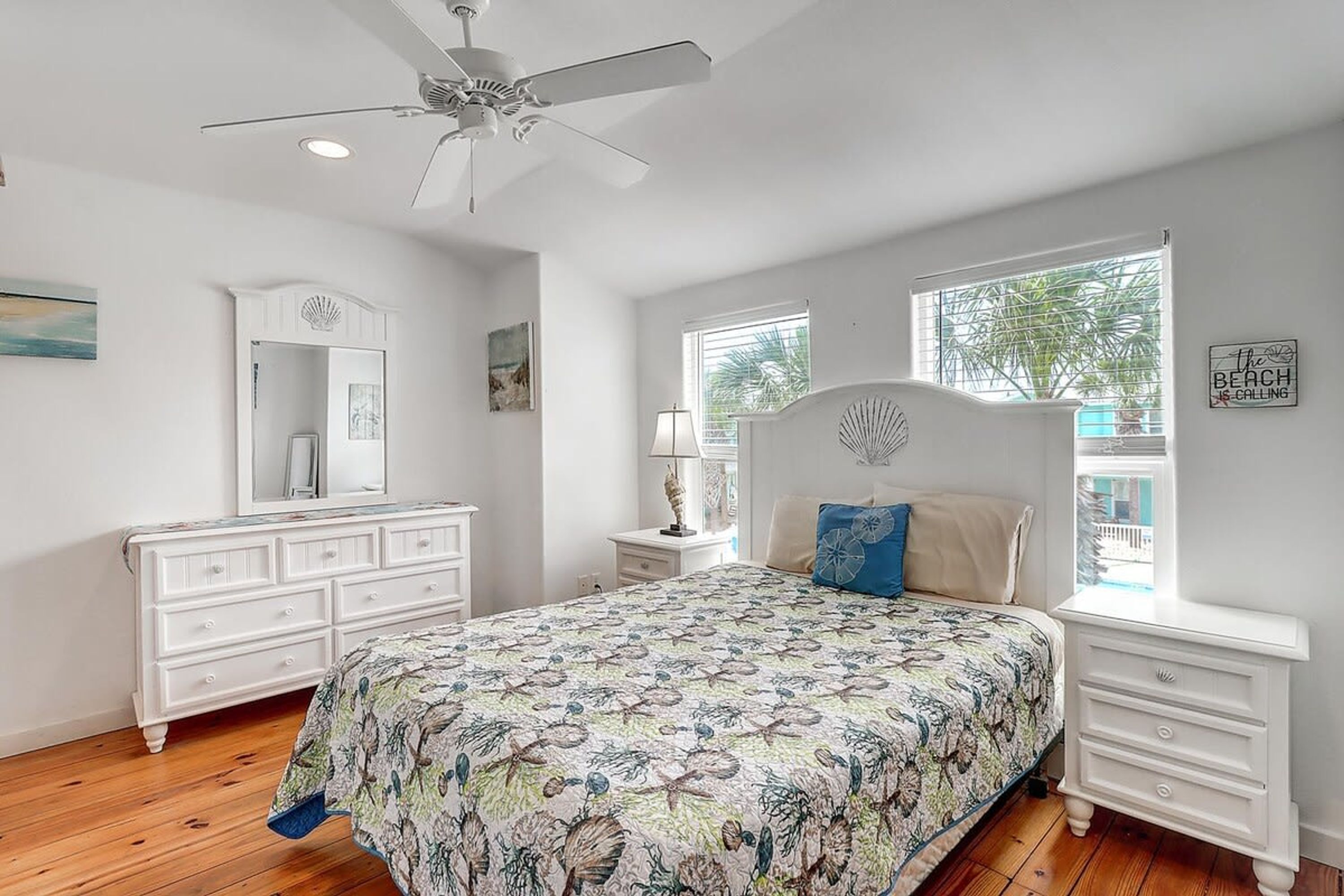 A bedroom with a bed, dresser, vanity, ceiling fan, and windows.