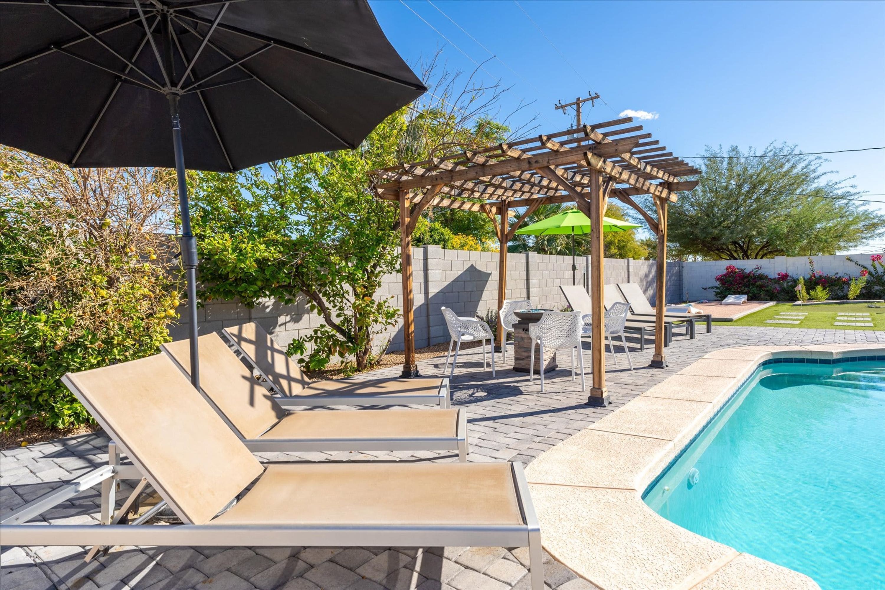 A backyard with a swimming pool, lounge chairs, a pergola, and a patio area.