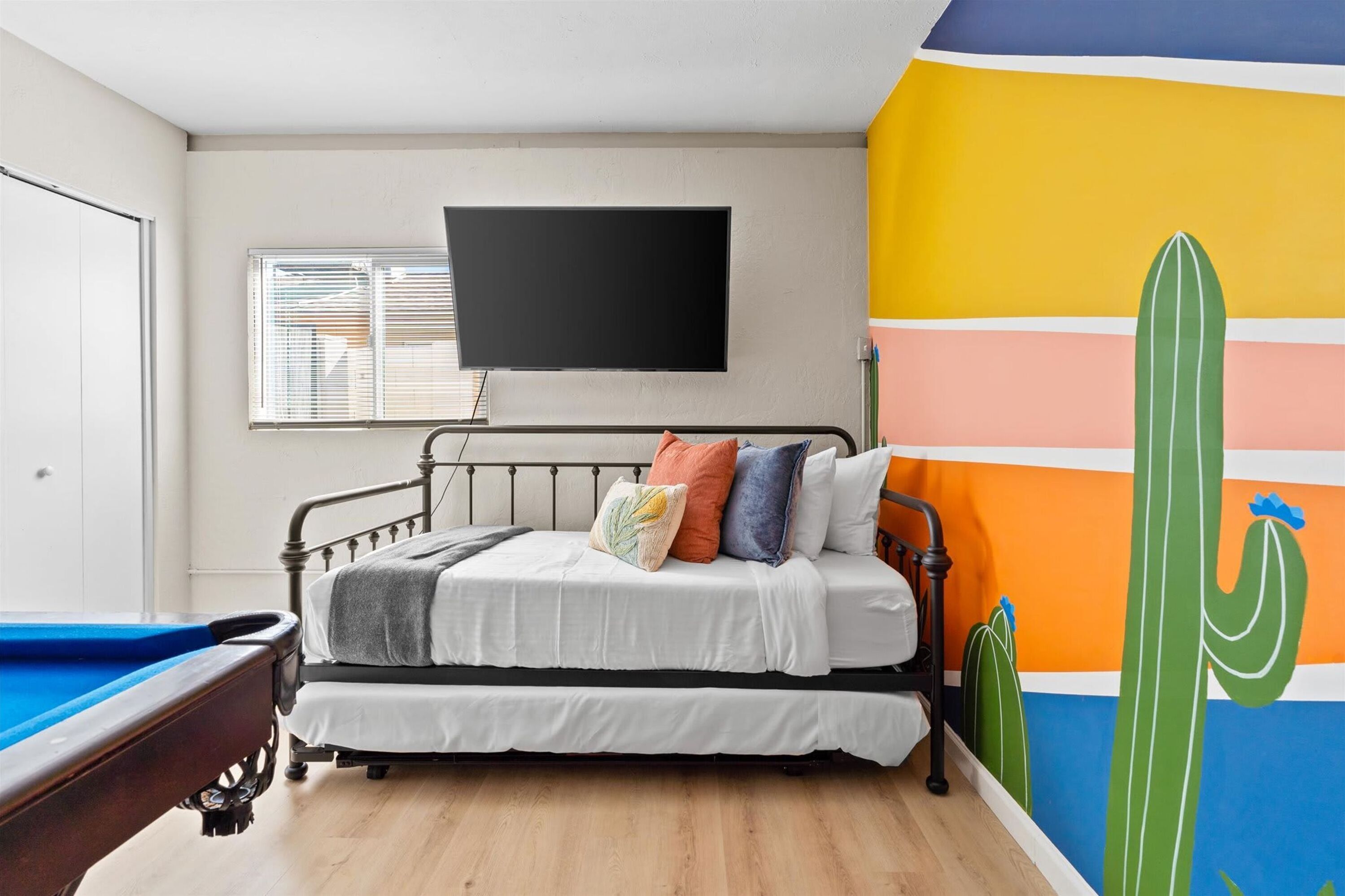 Een slaapkamer met een bed, een aan de muur bevestigde televisie, een raam met zonwering en een kleurrijke muurschildering.