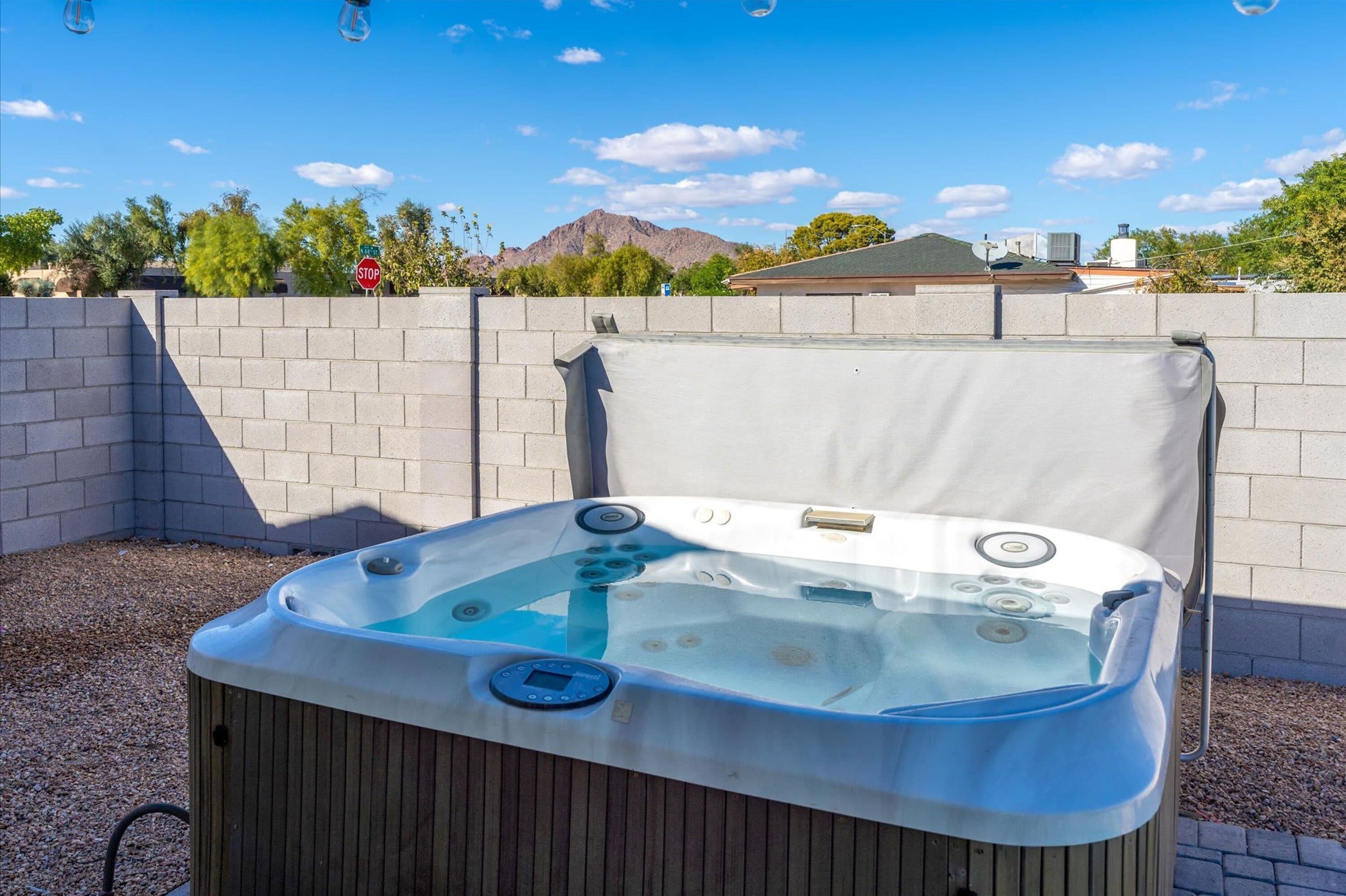 An outdoor hot tub with a control panel, surrounded by a brick wall and gravel, with a clear sky and mountains in the background.