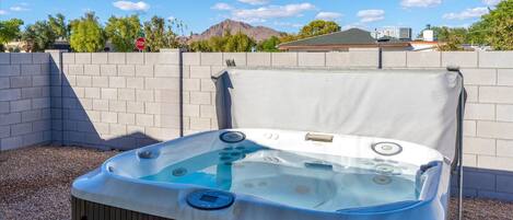 An outdoor hot tub with a control panel, surrounded by a brick wall and gravel, with a clear sky and mountains in the background.