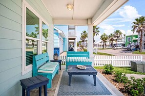 A porch with a blue bench and table, a white picket fence, and a view of palm trees and houses. - Island Gem- 4 BR Golf Cart Accessible Boardwalk (Port Aransas)