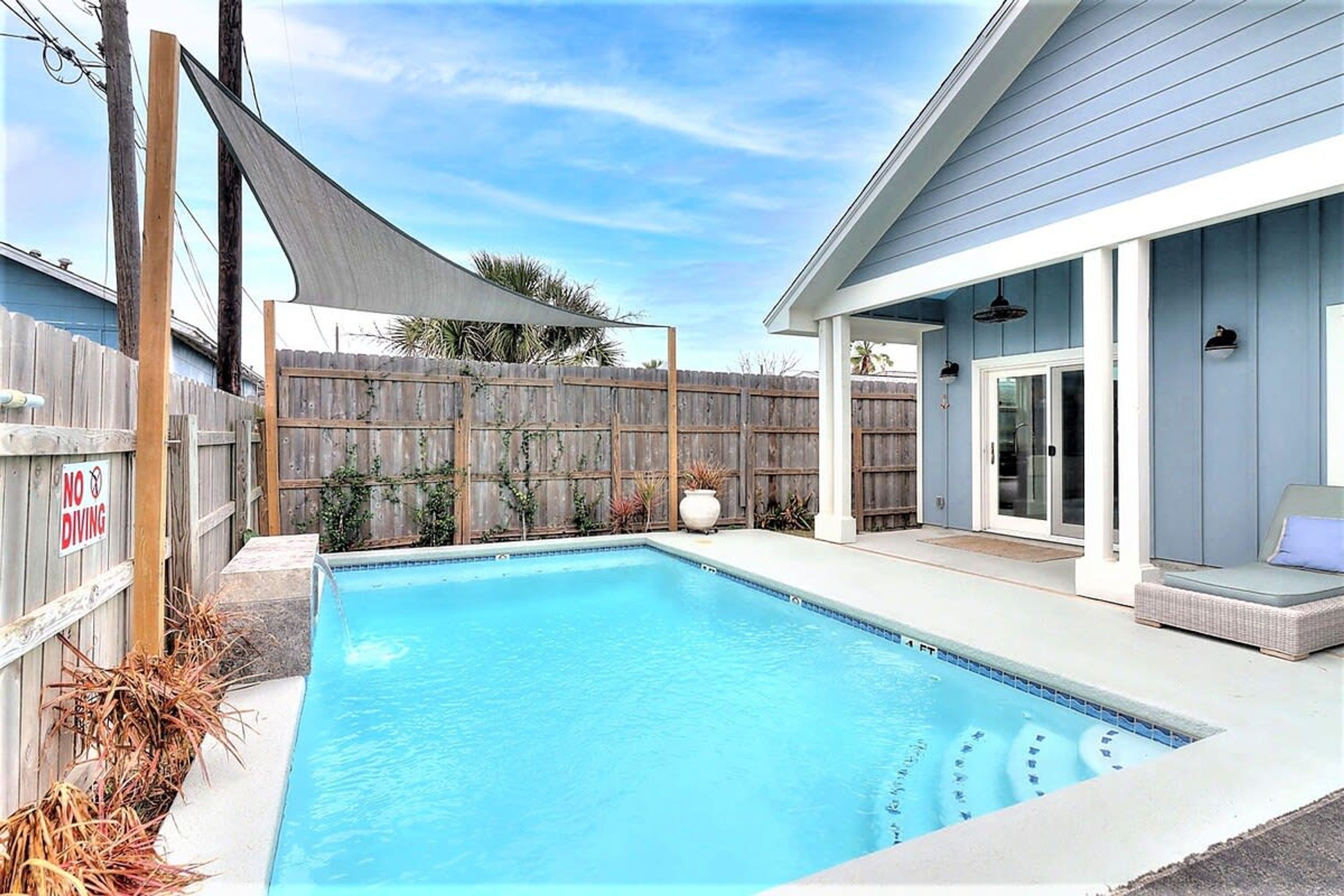 A swimming pool with a wooden fence, a shaded area, and a covered patio with a chair.