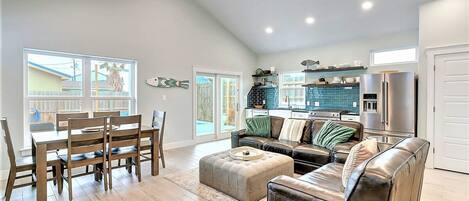 A spacious living room with a dining area, a kitchen with a blue backsplash, and a large window providing natural light.
