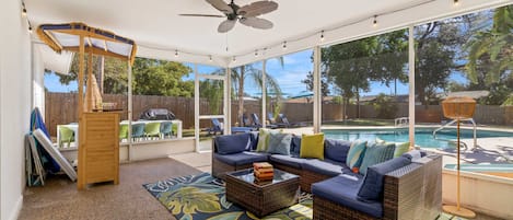 A covered outdoor seating area with a pool, wicker furniture, and a ceiling fan.