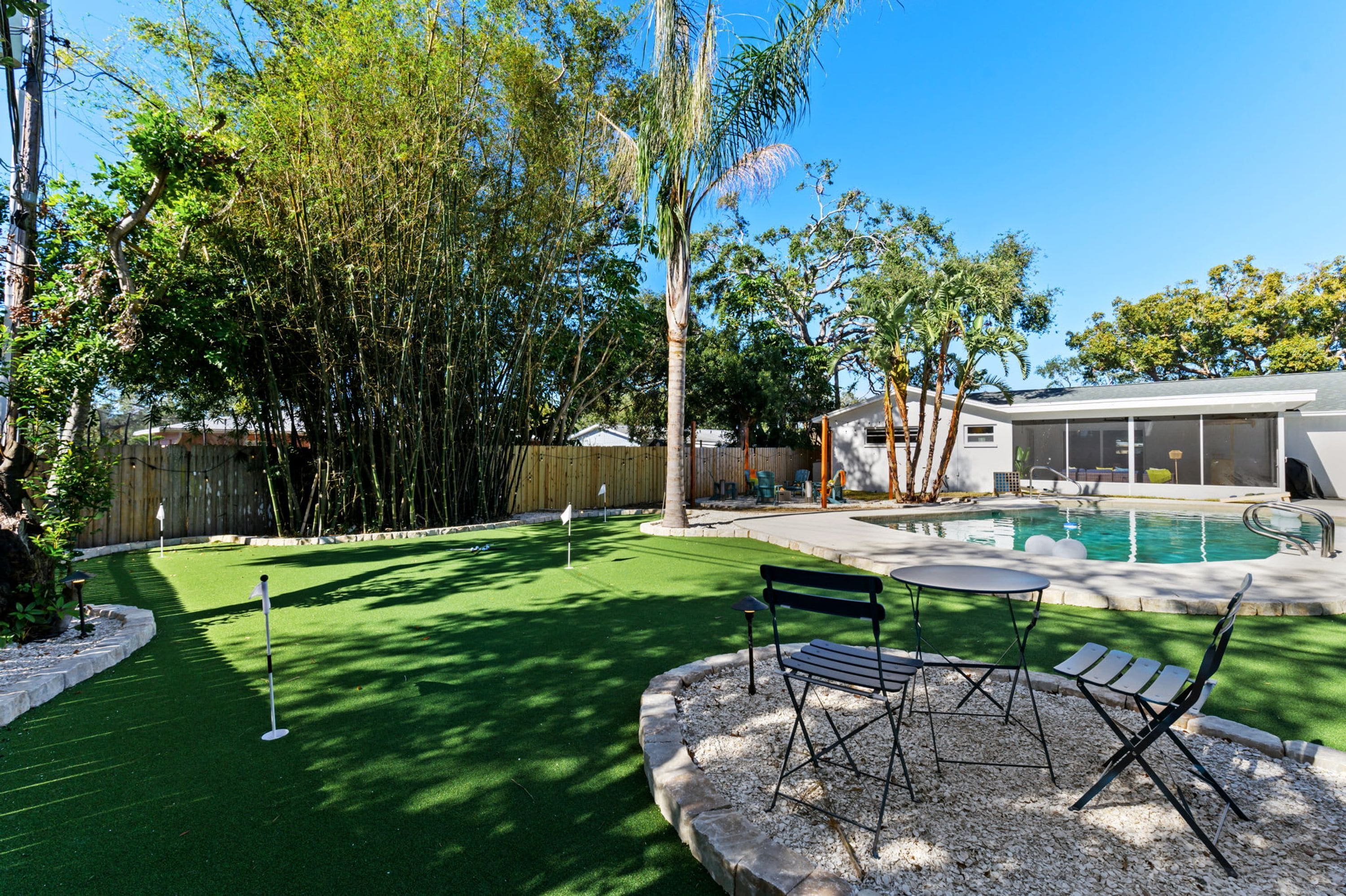 A backyard with a swimming pool, artificial grass, a patio with a table and chairs, and a wooden fence.