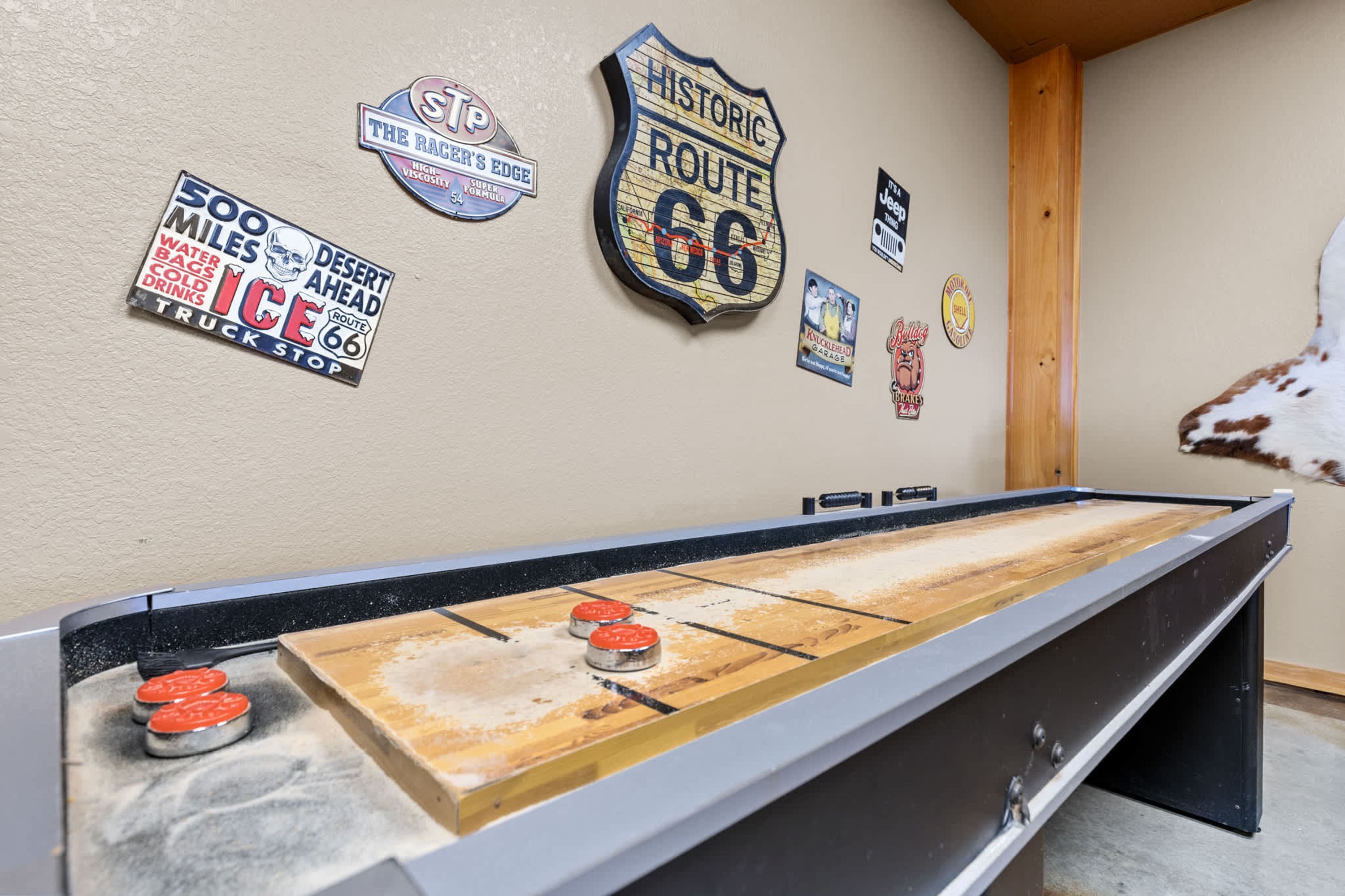 A shuffleboard table with a wooden surface and metal ends, surrounded by various wall decorations including a Route 66 sign and a desert-themed sign.