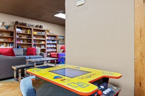 A room with a gaming console, a sofa, a coffee table, and a bookshelf filled with books and decorative items.