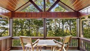 A covered outdoor seating area with a table and chairs, large windows, and a view of trees and hills. - Views for Miles Outdoor Fireplace Hot Tub (Morganton)