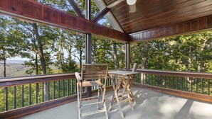 A covered outdoor area with a table and chairs, surrounded by a wooden railing, overlooking a forested landscape. - Views for Miles Outdoor Fireplace Hot Tub (Morganton)