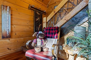 Interior acogedor de una cabaña con una escalera de madera, un sillón mullido con un oso de peluche y un estante de pared con objetos decorativos.