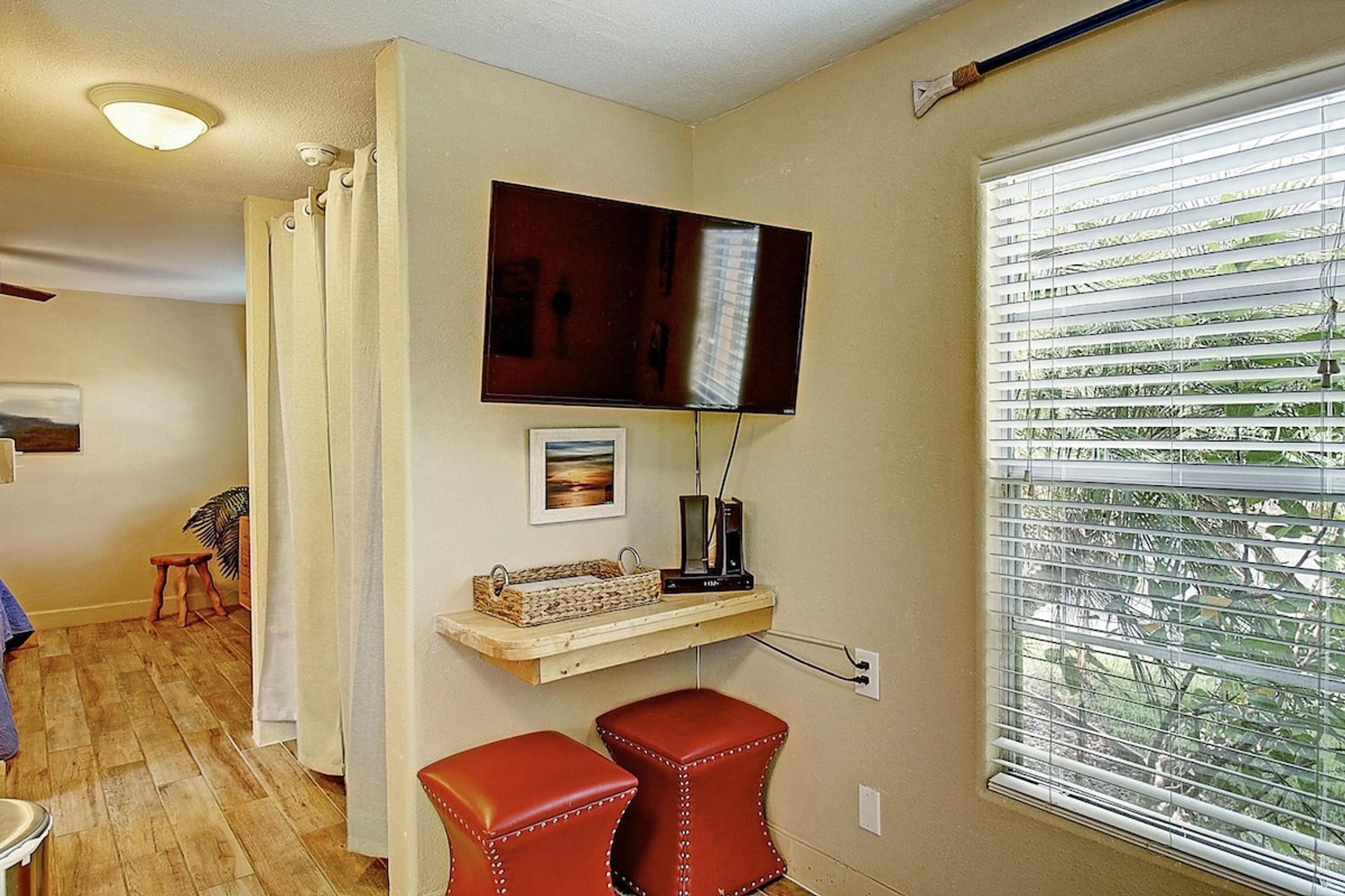 A room with a television mounted on the wall, a window with blinds, a small stool, and a shelf with a basket.