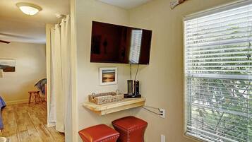 A room with a television mounted on the wall, a window with blinds, a small stool, and a shelf with a basket.