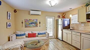A compact living space with a kitchenette, a sofa with cushions, a glass coffee table, and a painting on the wall.