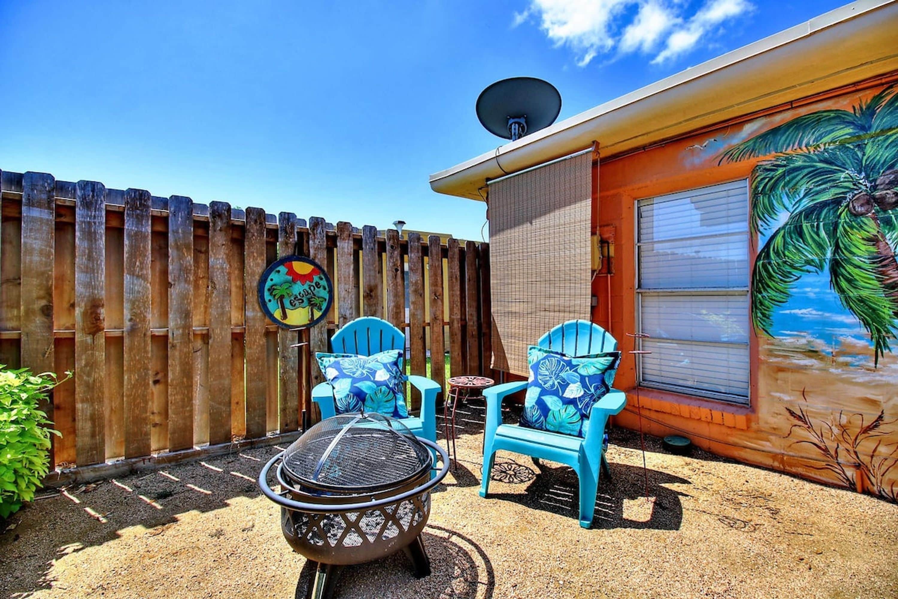 A backyard with a barbecue grill, two blue chairs, a satellite dish, and a palm tree mural on the wall.