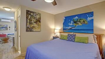 A bedroom with a bed, a ceiling fan, a painting of a palm tree, and a view of the ocean.