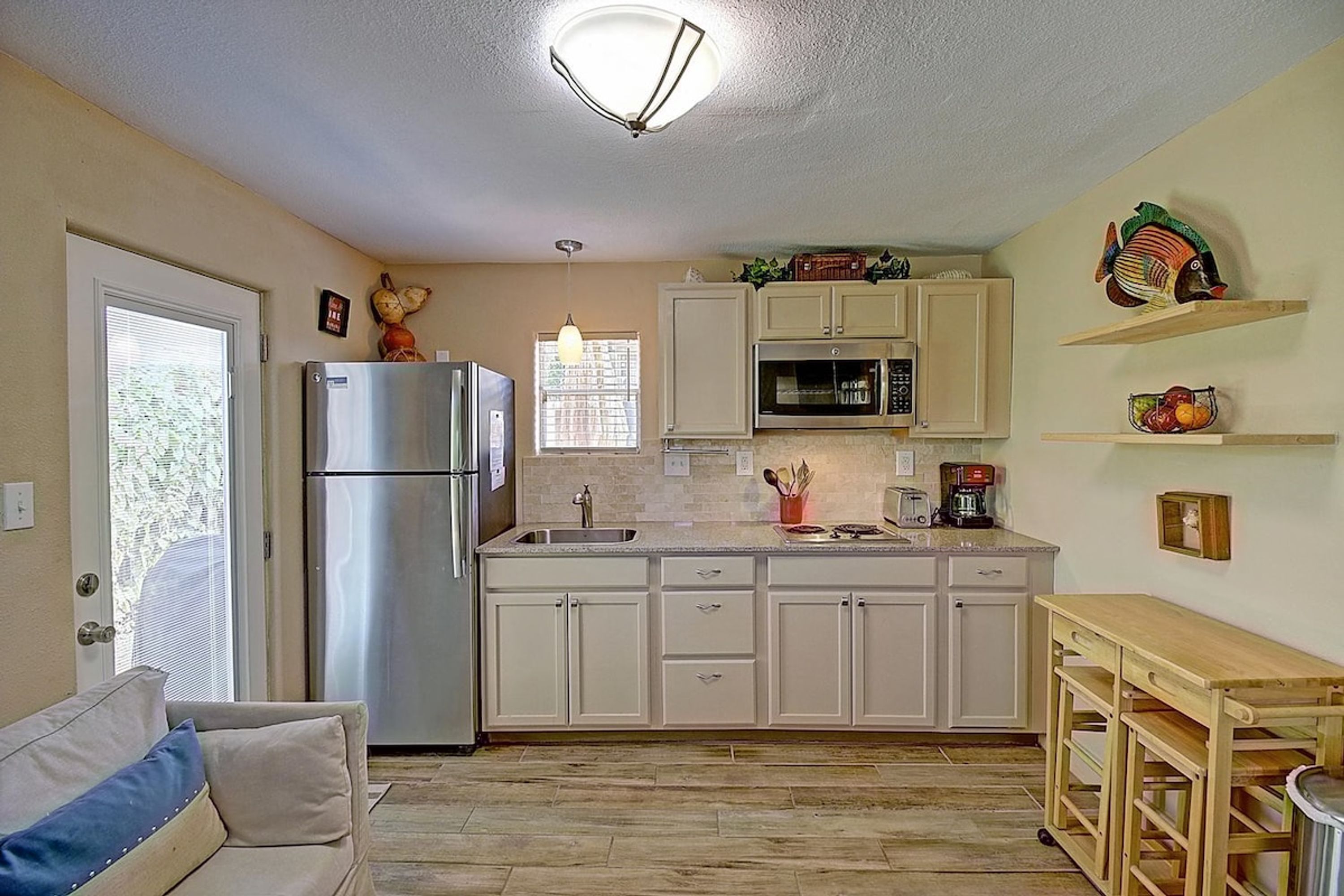 A compact kitchen with a refrigerator, microwave, and a sink. There is a wooden table and a chair, and a small shelf with decorative items.