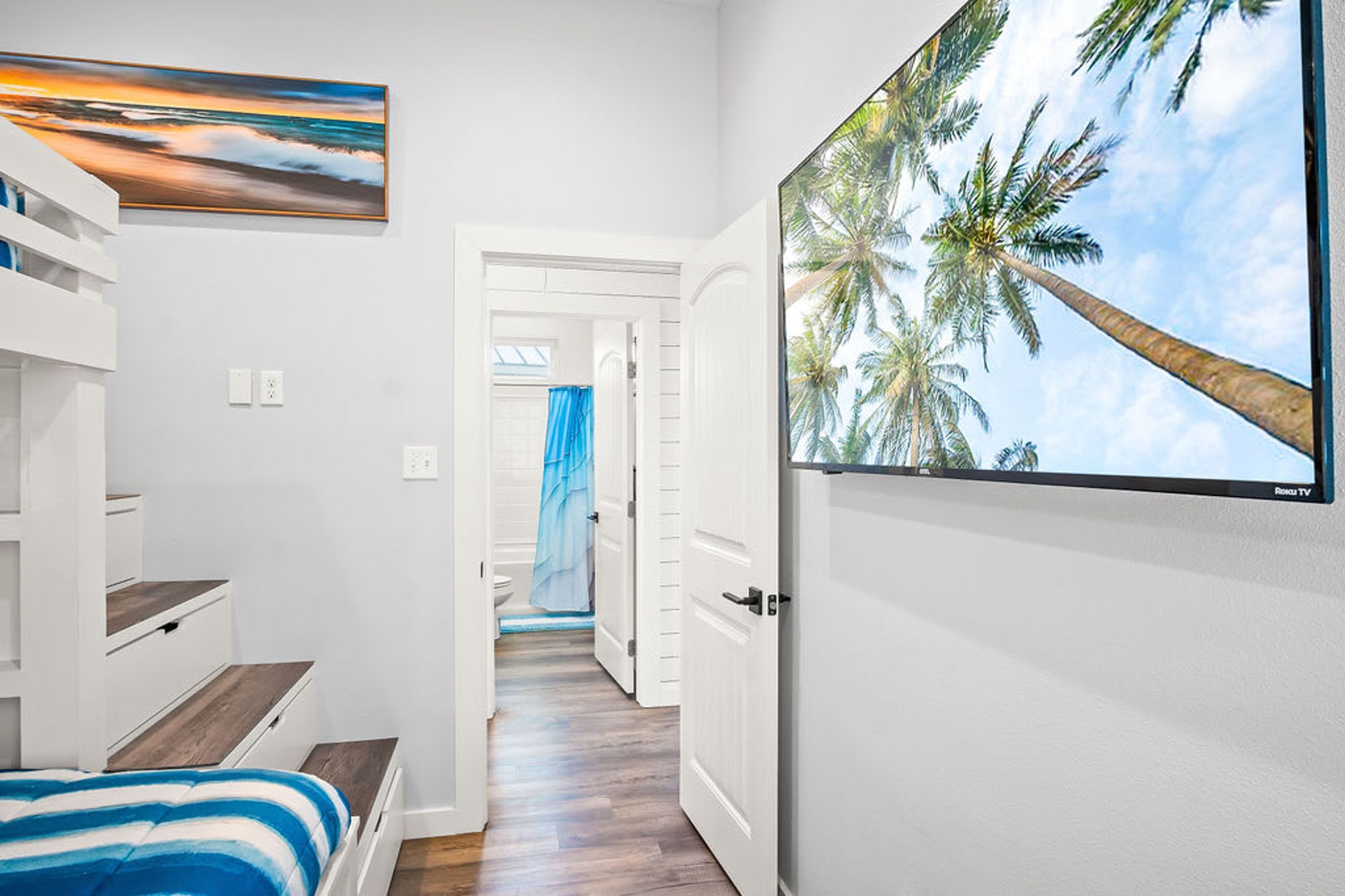A hotel room with a bed, a painting of a sunset, a large TV screen displaying palm trees, and a view of the hallway.