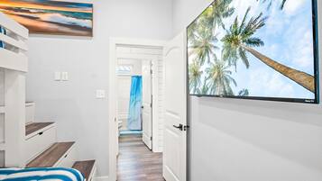 A hotel room with a bed, a painting of a sunset, a large TV screen displaying palm trees, and a view of the hallway.