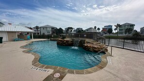 A swimming pool with a waterfall feature, surrounded by a stone deck and a metal fence. - Mustang Island Beach Side Sanctuary Game Room (Port Aransas)