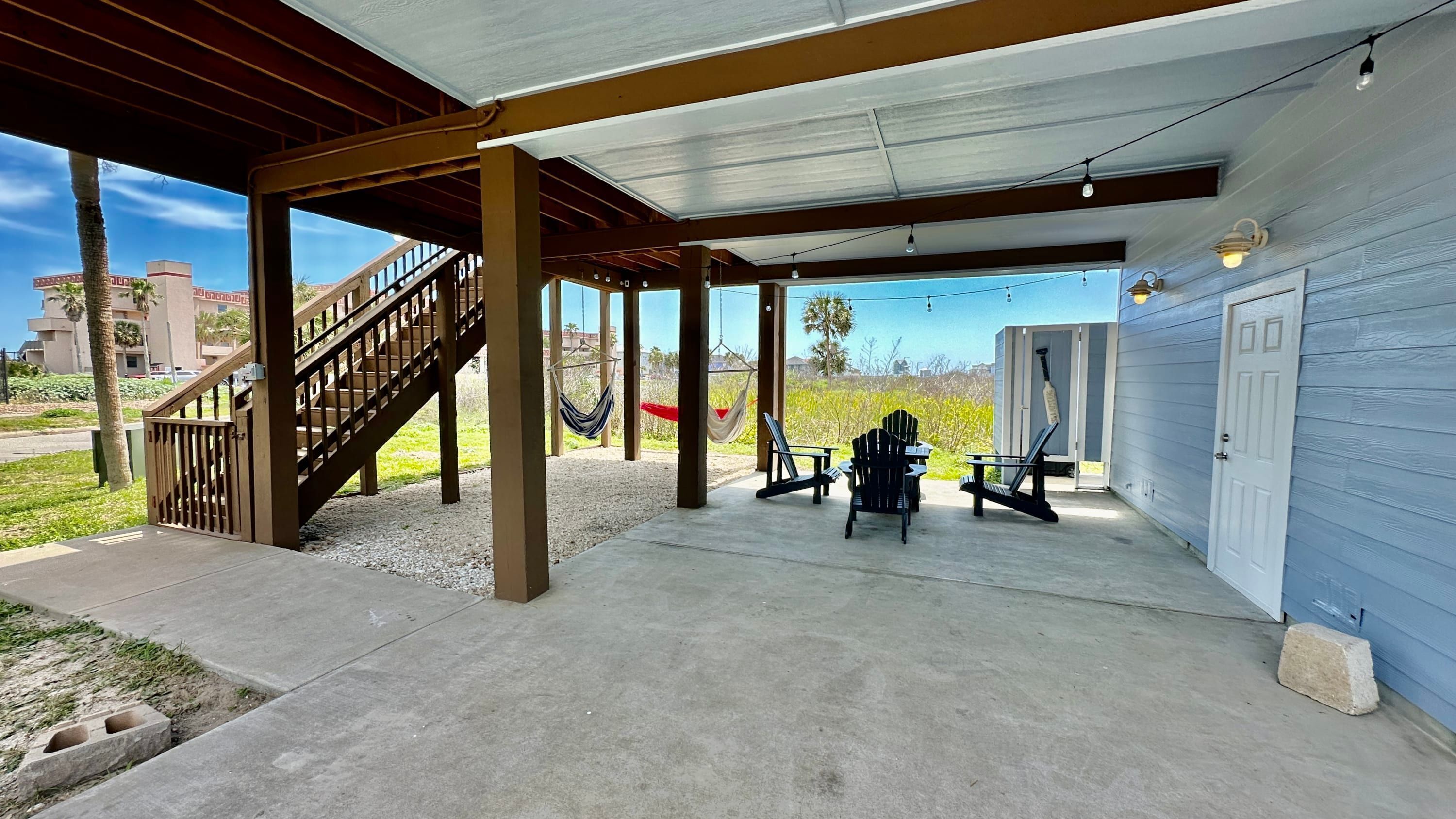 Un espacio al aire libre cubierto con piso de concreto, pilares de madera, una escalera y mobiliario exterior.