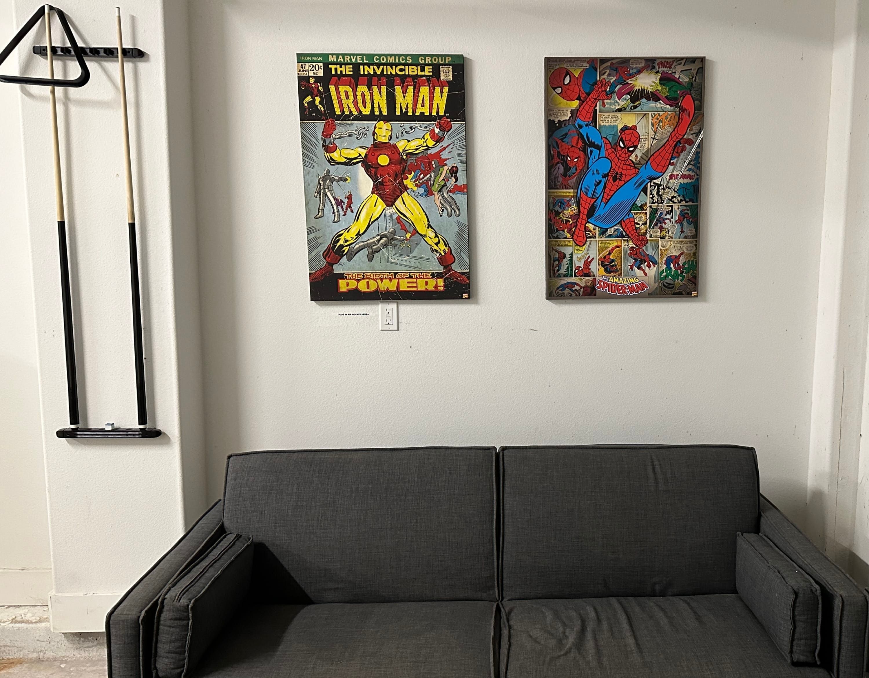 A room with a grey sofa, two comic book posters on the wall, and a pool table with cues.