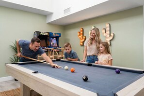 A pool table with four people around it, a children's game machine, and a cactus decoration on the wall. - The Sunset Peak Sleeps 18 Heated Pool Hot Tub (Phoenix)