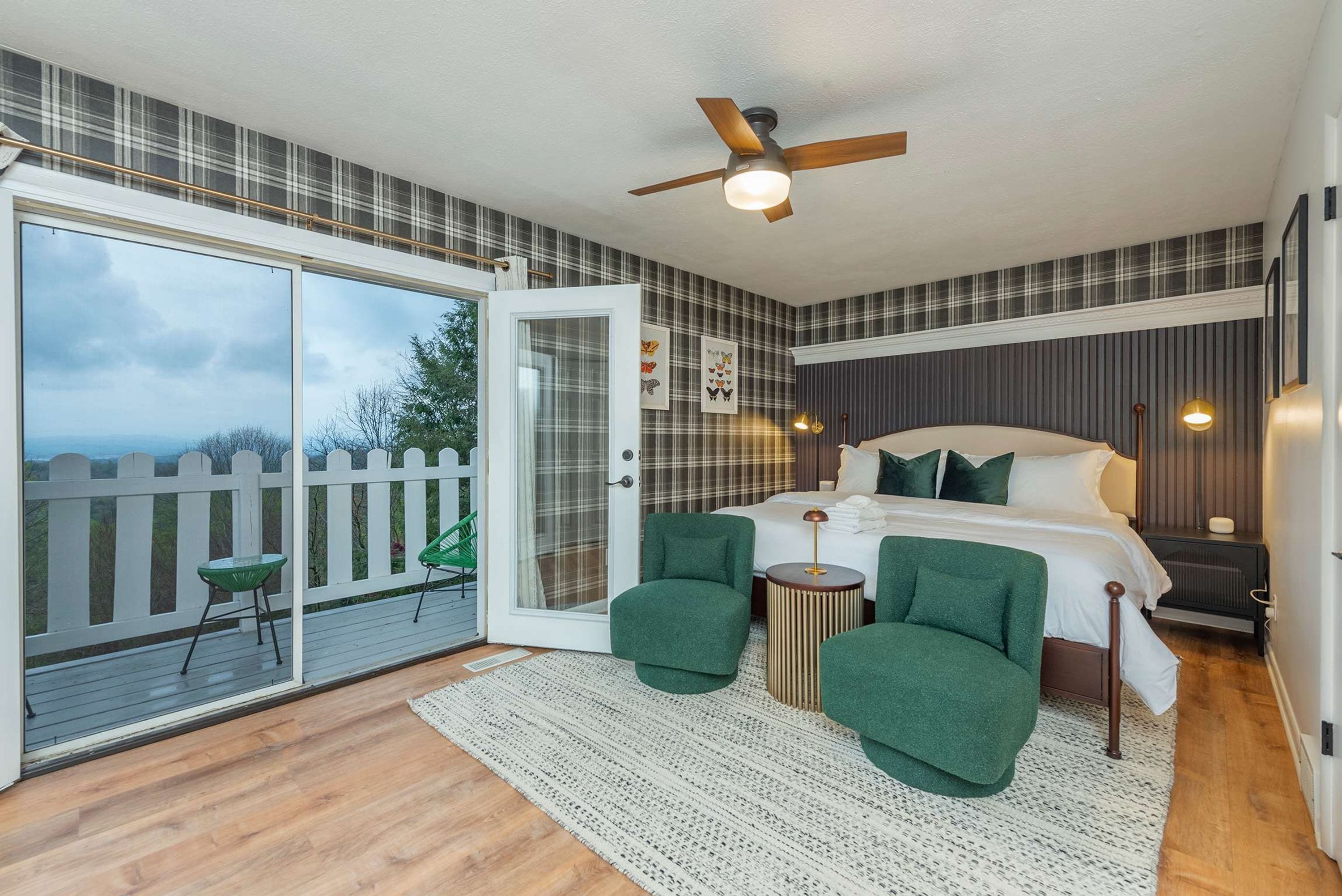 A bedroom with a bed, two green chairs, a round side table, a ceiling fan, and a view through a sliding glass door to an outdoor deck.