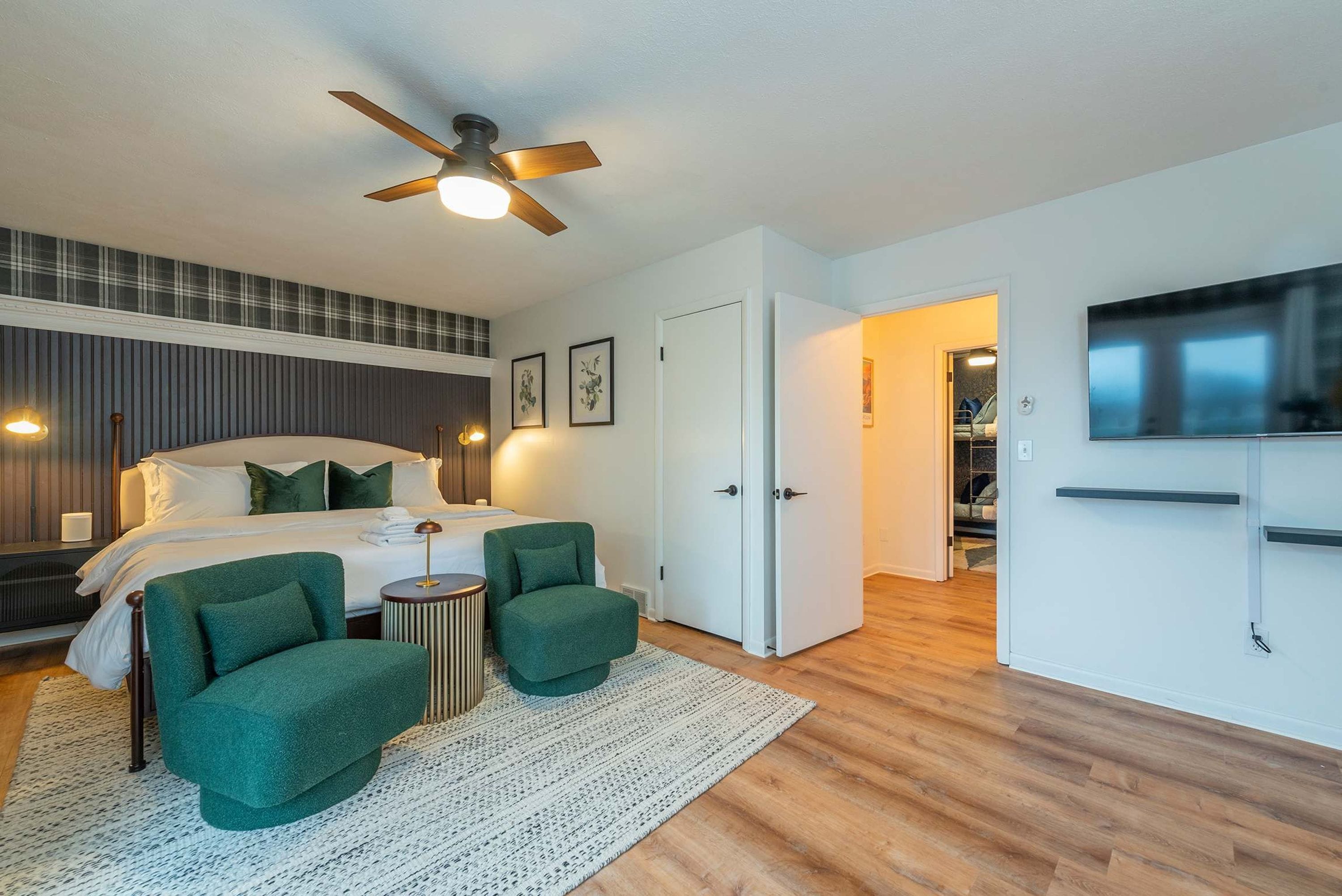 A modern bedroom with a bed, two green armchairs, a ceiling fan, and a TV mounted on the wall.