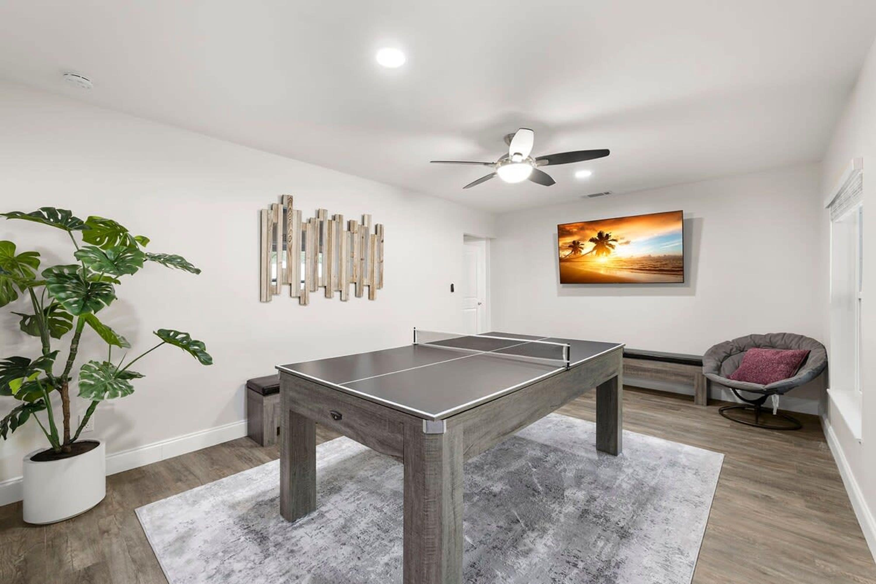 Una habitación con mesa de ping pong, ventilador de techo, un cuadro de un atardecer y una planta en maceta.