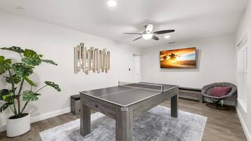 Una habitación con mesa de ping pong, ventilador de techo, un cuadro de un atardecer y una planta en maceta.