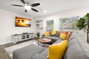 Un salon avec un canapé gris, une table basse, un ventilateur de plafond et des œuvres d’art accrochées au mur.