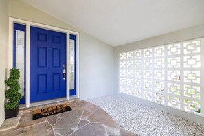 Een blauwe deur met een deurmat, een witte muur en een decoratieve witte scherm met geometrische patronen.