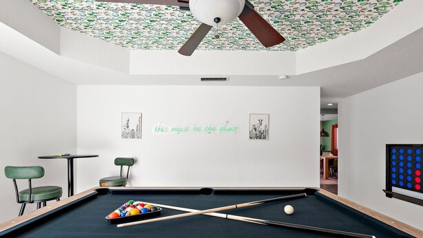 Ein Billardtisch mit Kugeln und Spielstöcken, ein Deckenventilator und eine Wand mit Kakteen-Kunstwerk.