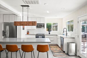 Une cuisine moderne avec des meubles blancs, des appareils électroménagers en acier inoxydable, un îlot central doté de tabourets et une vue sur l’extérieur grâce à de grandes fenêtres.