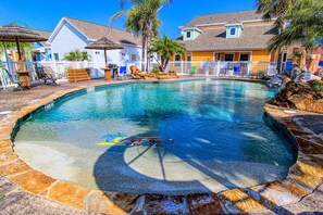 Una piscina con borde de piedra, rodeada de casas con vallas blancas y palmeras.
